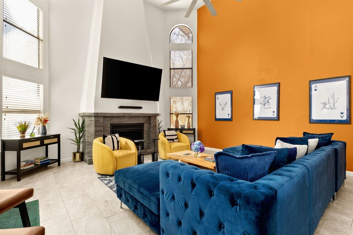 Sun-soaked living room with soaring windows, bold accent wall, plush blue sectional, cozy yellow chairs  and big-screen TV—perfect for movie nights, gathering with friends, and unwinding after exploring San Antonio.
