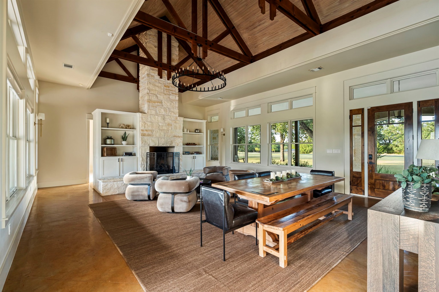 A spacious and inviting living area featuring high ceilings with exposed beams, a large dining table with benches, a cozy fireplace, and ample natural light.