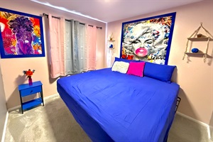 The bedroom exudes vibrant & eclectic energy with its bold, colorful artwork & furnishings. The large bed, adorned with deep blue linens, invites relaxation, while the bright artwork adds a playful touch. Natural light streams through the window.
