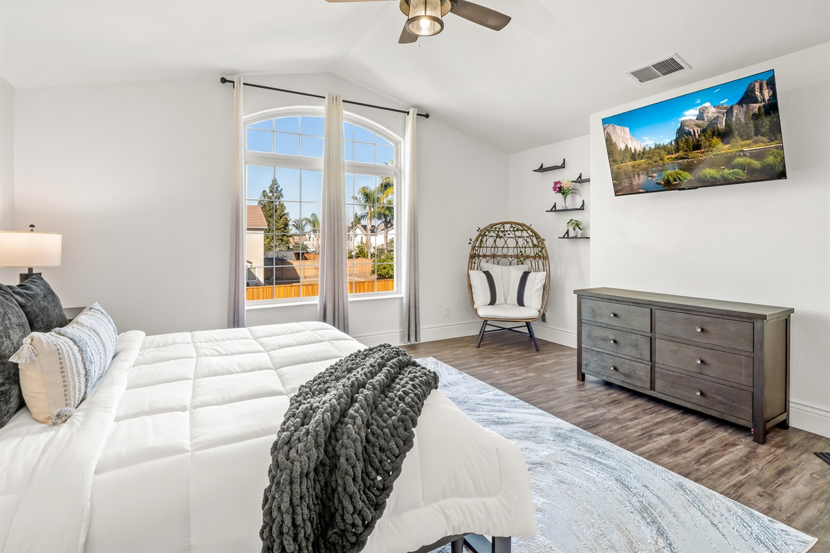 Elegant primary retreat featuring dramatic vaulted ceilings, a statement window, stylish seating nook, and in-room TV—luxury, light, and comfort all in one.