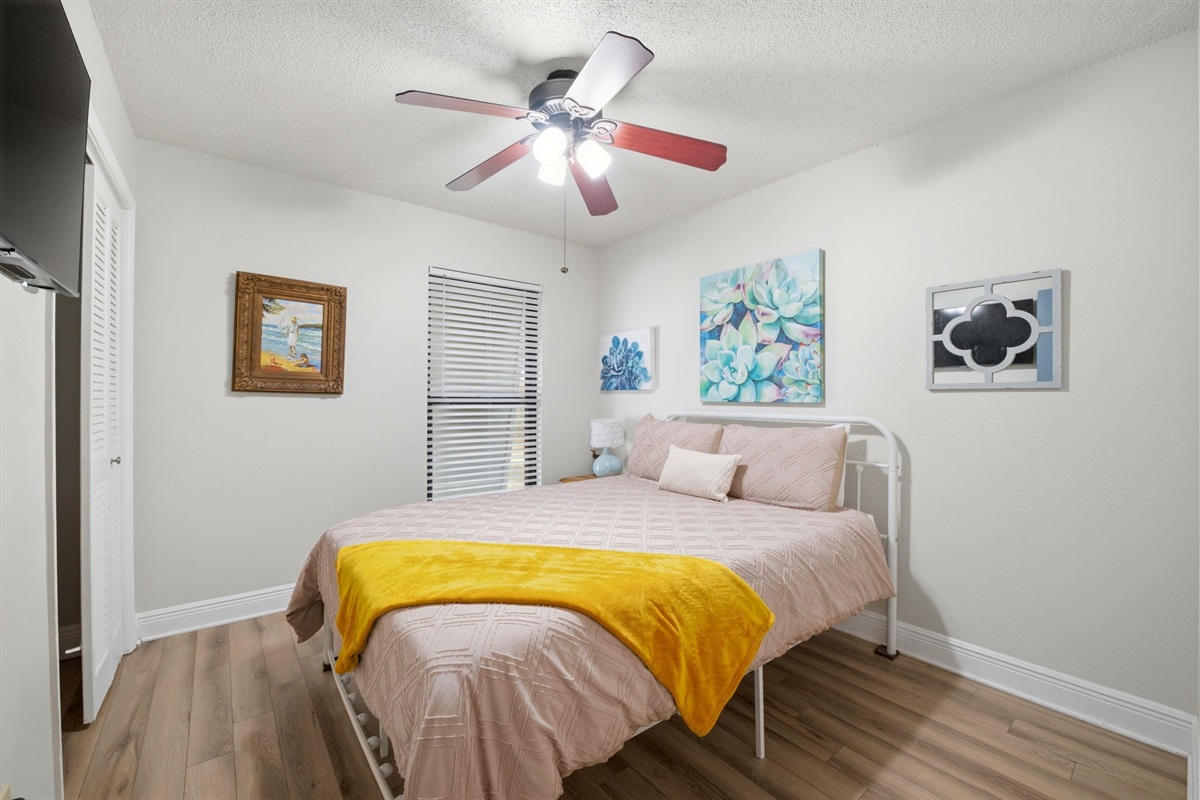 This charming bedroom features a comfortable bed, colorful coastal artwork, and a ceiling fan for added comfort. Soft tones and natural light create a bright and relaxing space, perfect for unwinding after a day at the beach.