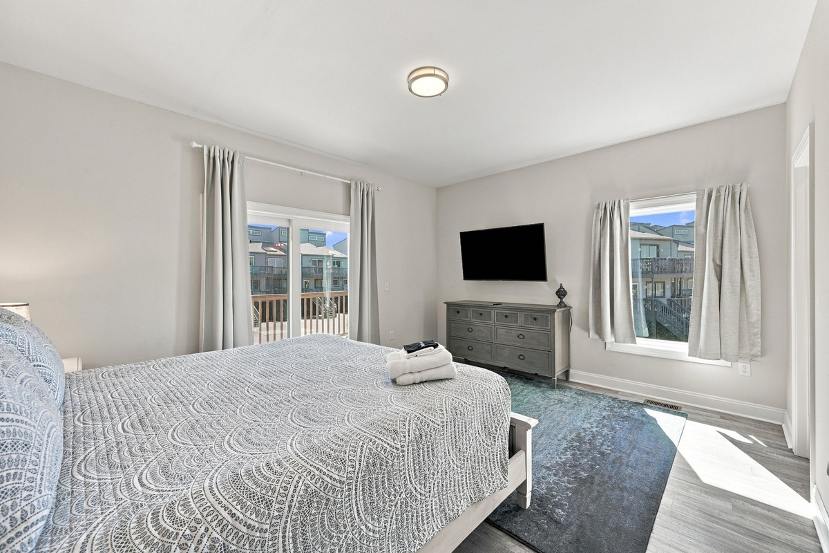 Serene view of the third bedroom highlighting a wall-mounted flat-screen TV, dark wood dresser, teal area rug, and sliding glass doors to the private balcony.   