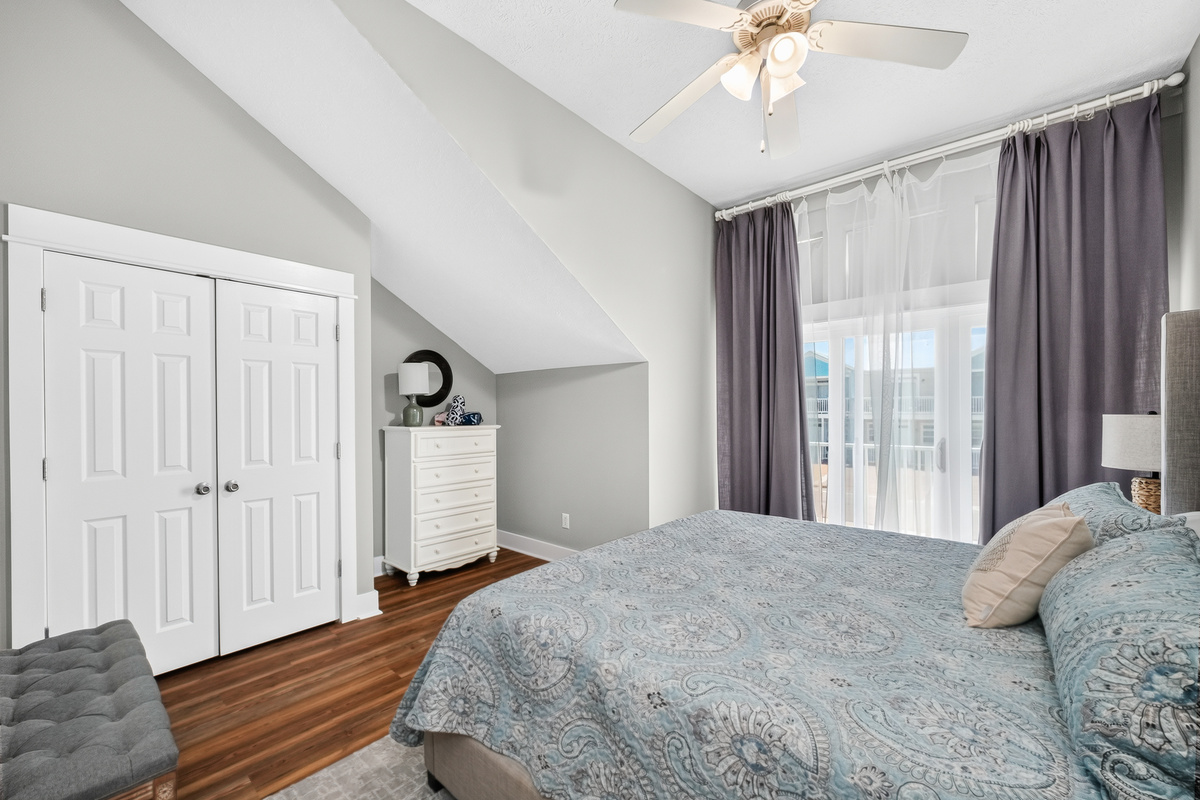 Comfortable king-size bed, plush linens, ceiling fan, spacious dresser, closet storage, bedside lamp, and a bright outdoor balcony.