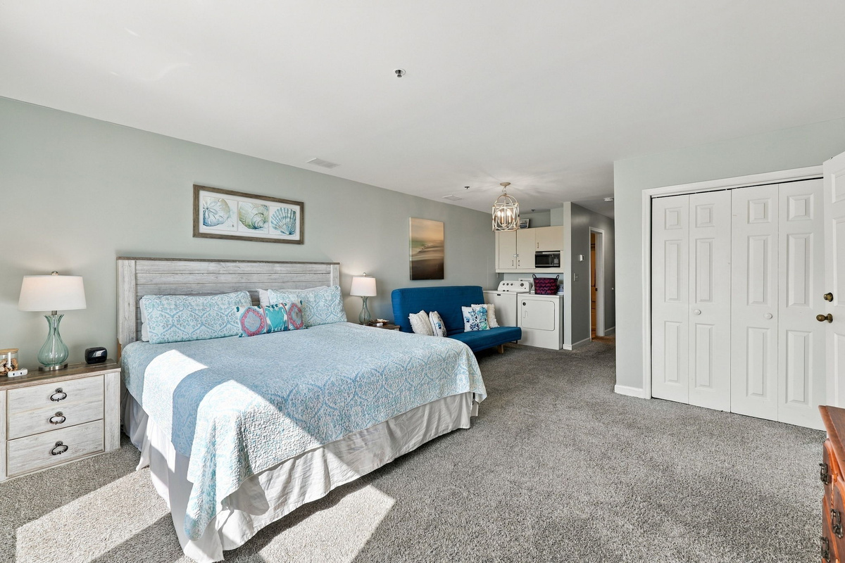 Serene primary bedroom retreat featuring a king bed with a weathered wood headboard, subtle coastal accents, spacious bi-fold closet storage, and integrated sitting/laundry area beyond.   