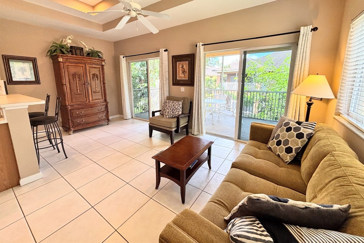Comfortable living area with high ceilings, tile flooring, cozy seating, and two sets of sliding glass doors that bring in natural light and lead out to the private screened balcony.