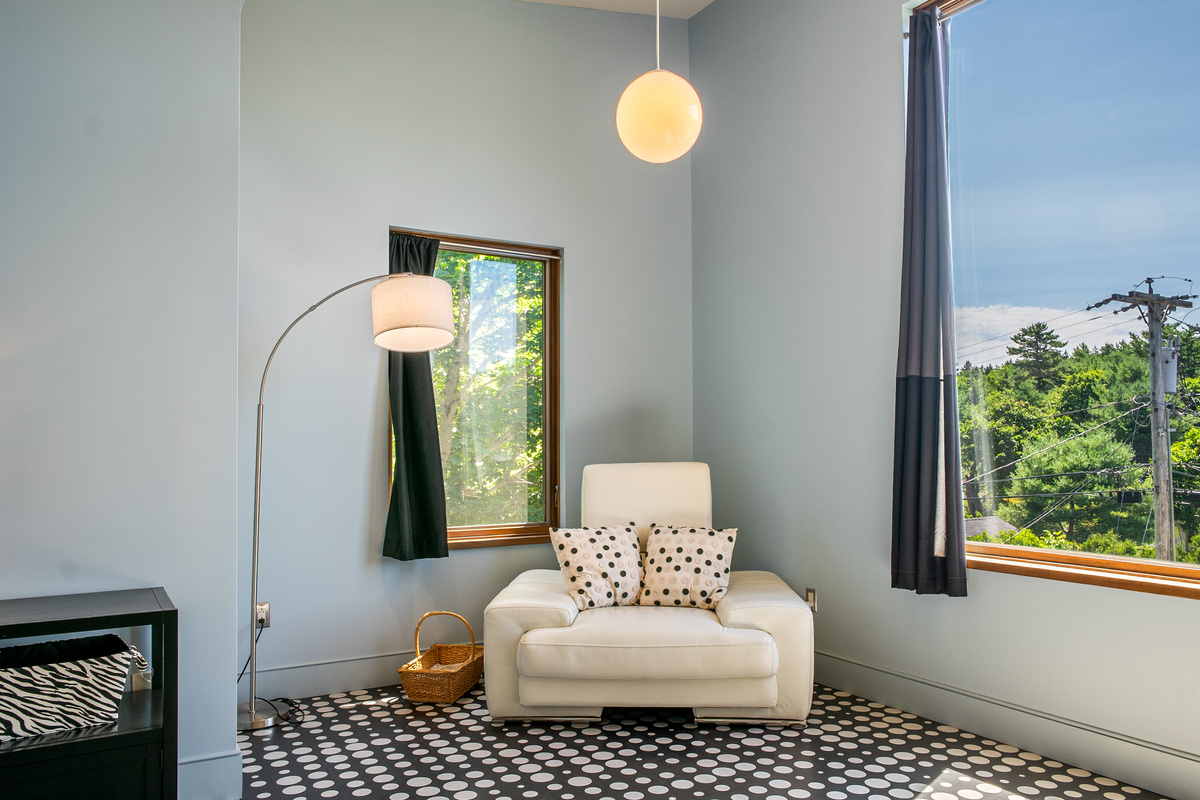 Sun-drenched serenity in "The Sky Loft" bedroom at "The Crag." Features soaring ceilings, a cozy oversized reading chair, stylish patterned flooring, and large picture windows framing tree-lined views for a peaceful top-floor retreat.