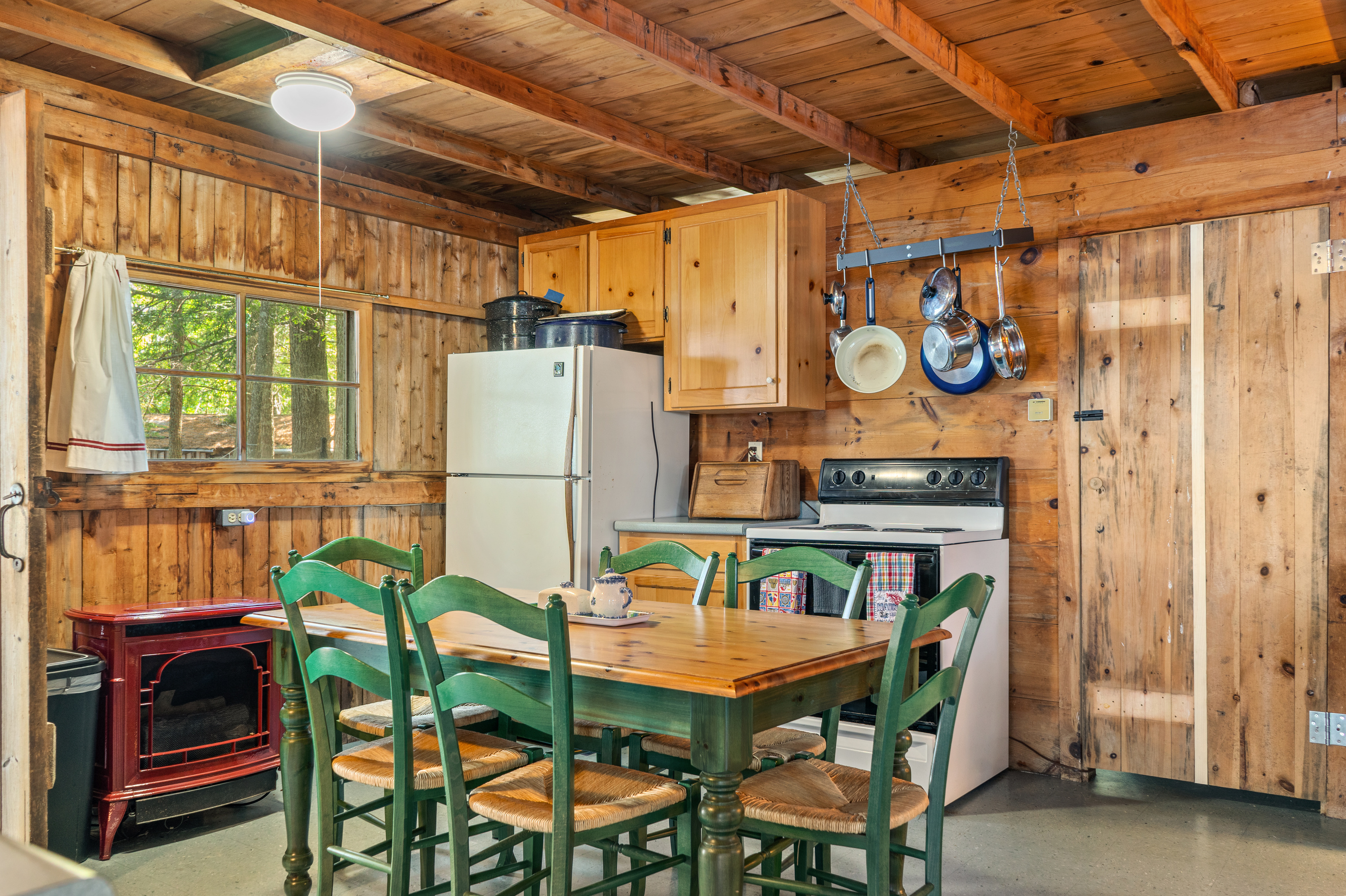 Dining area & primary heat source at Loonrise Point! Red heater stove provides cozy warmth for cool Maine days. Features full dining set for 6, full-size fridge, electric range, hanging pot rack, & knotty pine style on Beech Hill Pond in Otis, ME.