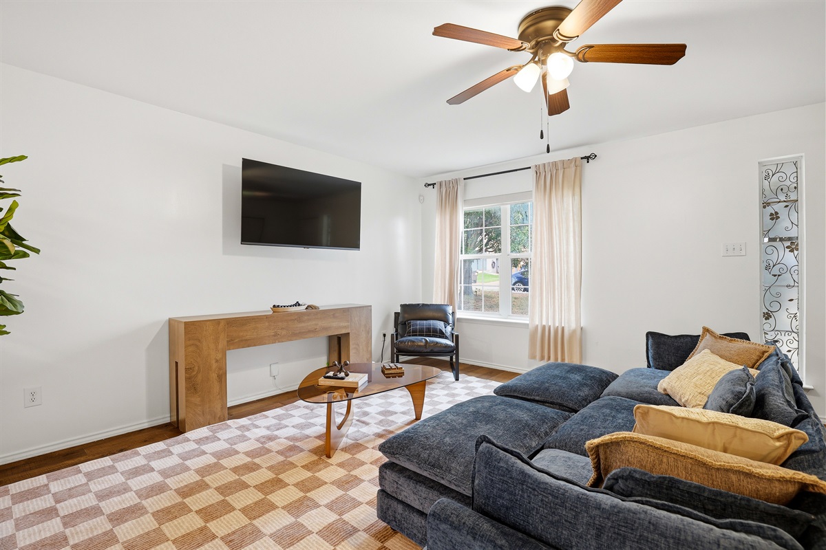 Spacious living area with oversized sectional sofa, wall-mounted Smart TV, ceiling fan, and checkered area rug. Open layout with natural light — great for relaxing or gathering as a group.