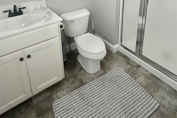 This bathroom features a white vanity with a marble-style top and dark hardware. The space includes a standard toilet, a walk-in shower with frosted glass doors, and grey tile flooring accented by a textured bathmat.