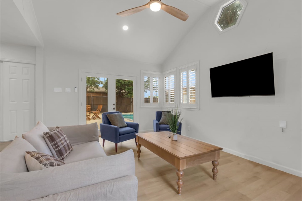 Relaxed living area with a mounted TV, bright windows, and direct access to the private entrance leading to the pool and backyard.
