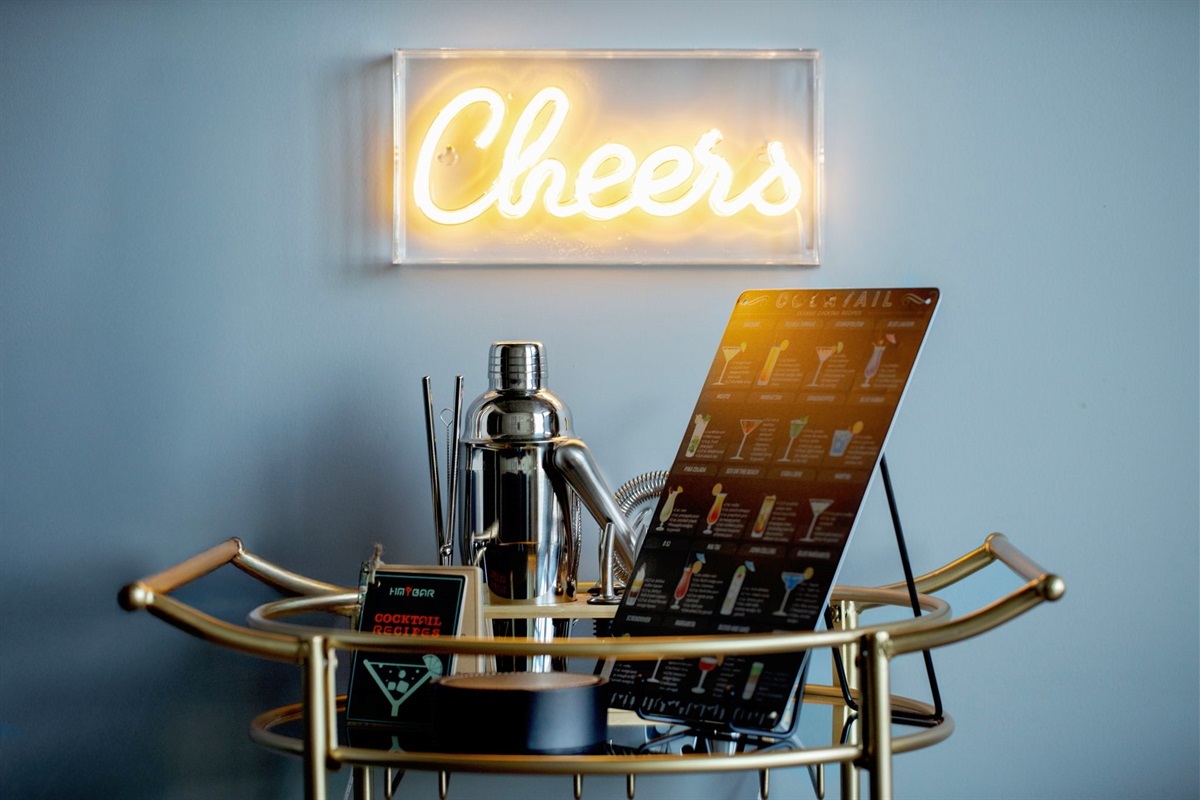 A gold bar cart styled with a cocktail shaker, mixing tools, and a guide to various drinks. A neon "Cheers" sign hangs on the gray wall above, creating a fun and inviting atmosphere.