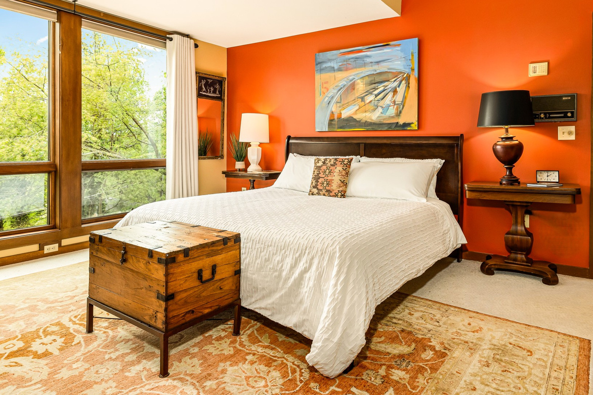 Main-floor primary suite featuring a premium Helix King bed, high ceilings, and large windows overlook the private, treed property. Decorated with vintage furniture and an antique area rug, just steps from the full en-suite bath.