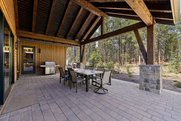 Covered outdoor dining and lounge space with a gas grill, surrounded by peaceful pine forest