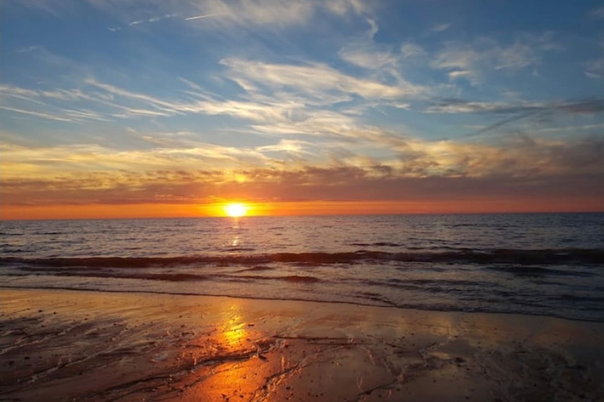 Take in the stunning horizon where the setting sun meets the ocean waves, casting a warm golden light across the wet sand. The wispy clouds add a dramatic touch to this tranquil coastal scene, located just a short walk from your front door.