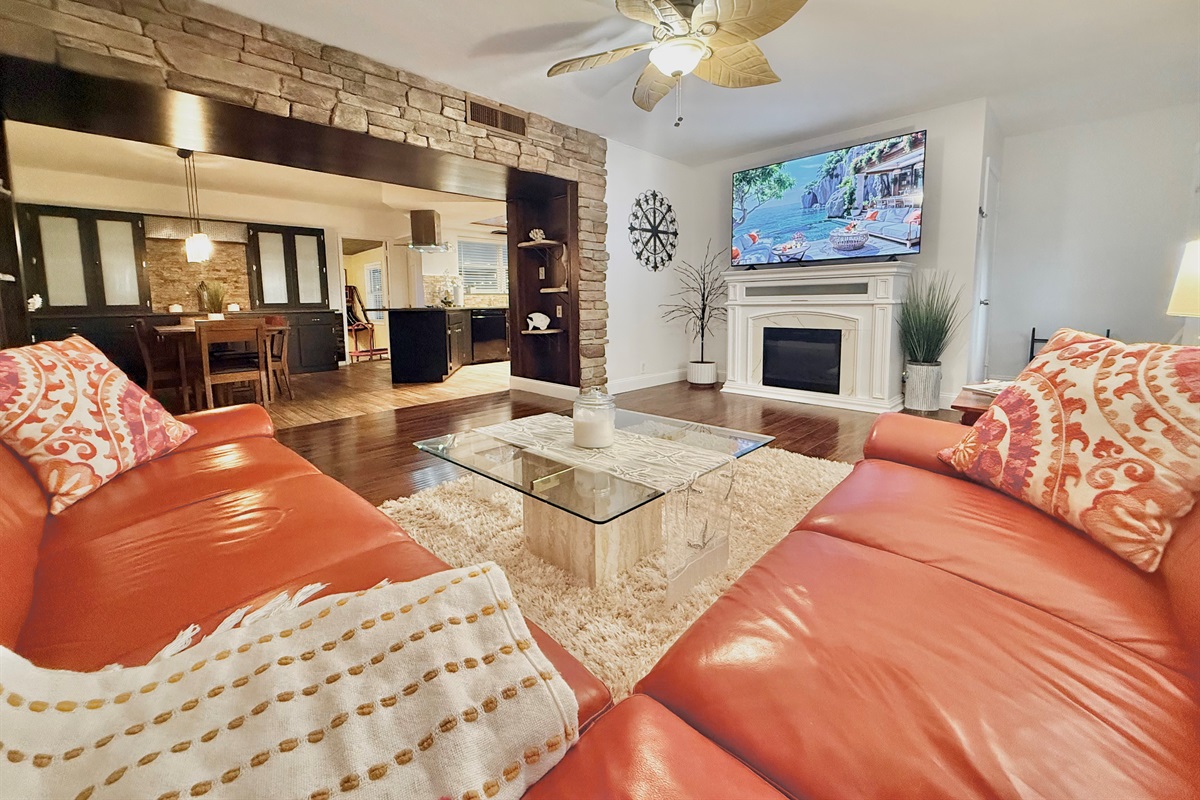 This inviting living room features a large wraparound sofa, warm wood floors, and a smart TV over a charming fireplace—perfect for movie nights or game-day lounging.