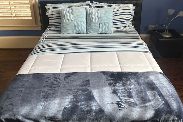 Cozy Bedroom 3 with a calming blue accent wall, dark wood floors, and a comfortable bed dressed in crisp striped bedding. Features a modern headboard, bedside table with lamp, and window with blinds for natural light.