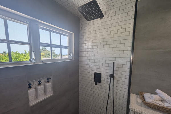 Walk-in shower at Villa Coral showcasing a matte black square rainfall showerhead, a matching handheld sprayer wand, white subway tiles, and built-in toiletry dispensers under bright windows.
