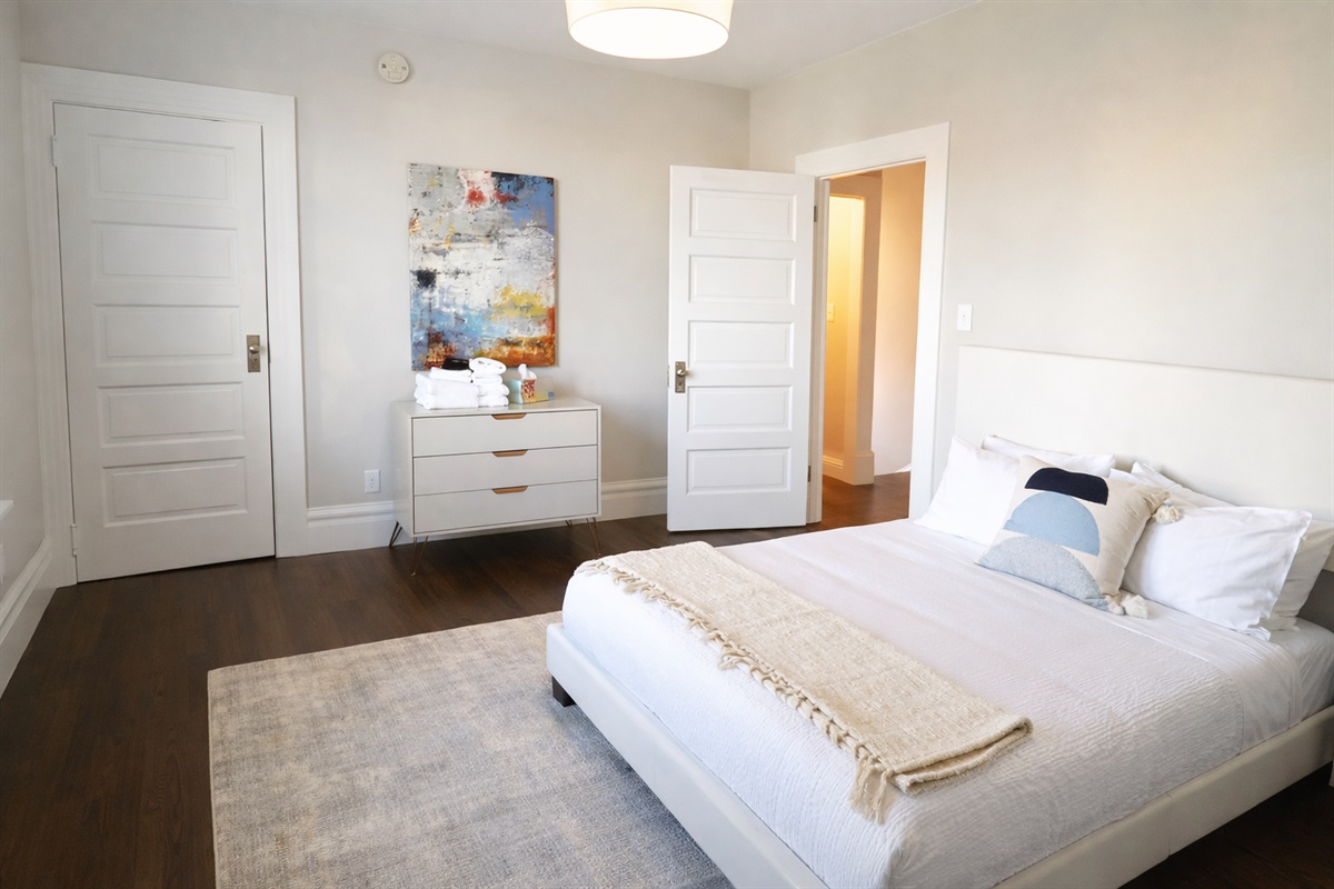 Retreat to the sun-drenched primary bedroom, featuring a comfortable king-size bed, designer art, and warm hardwood floors.
