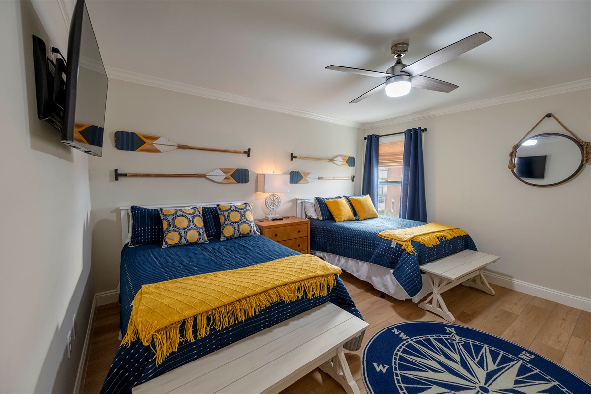 A cozy nautical-themed bedroom featuring twin beds with blue and yellow accents, wooden oars as decor, and a compass rug, creating a seaside-inspired atmosphere.