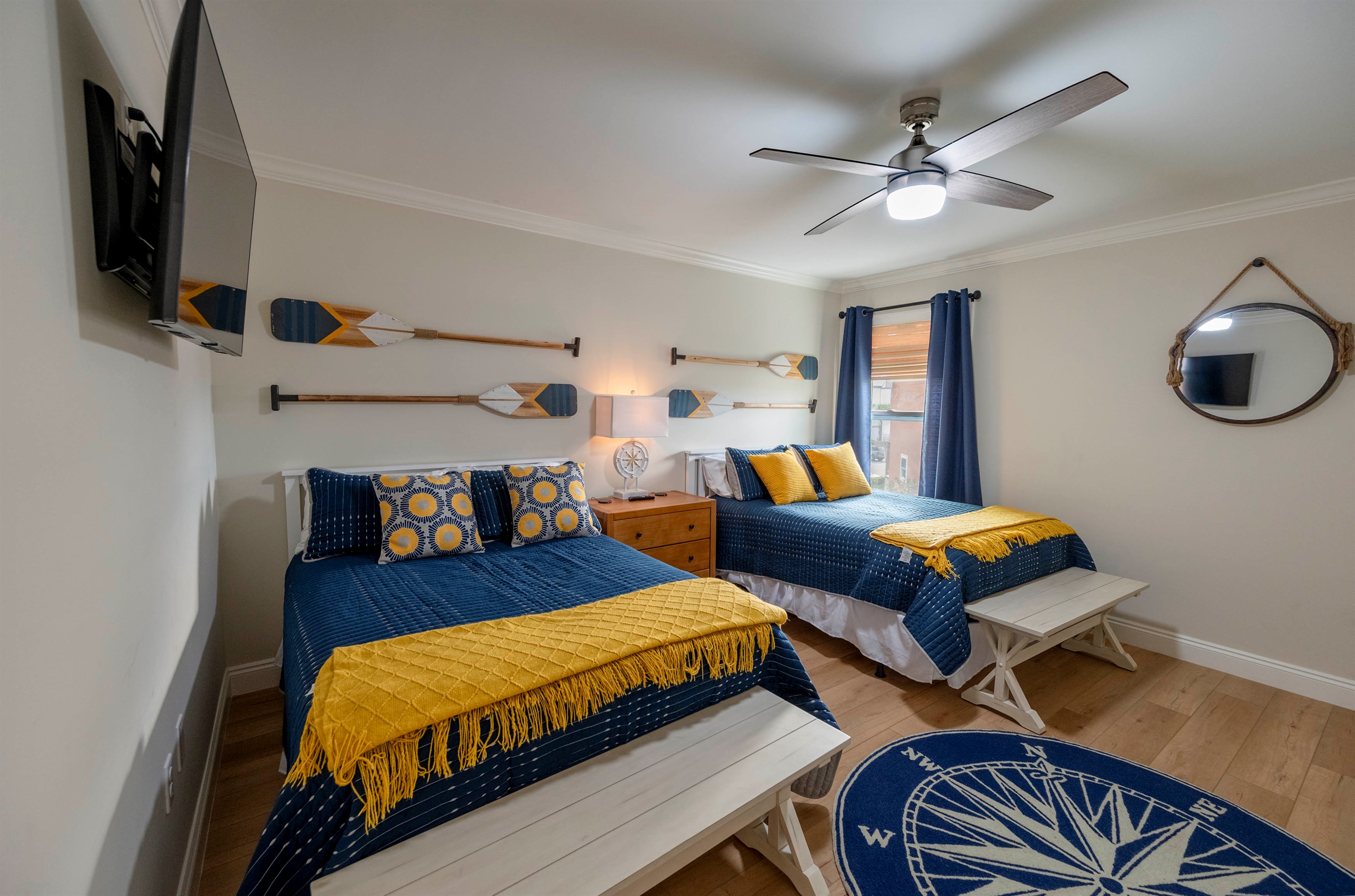 A cozy nautical-themed bedroom featuring twin beds with blue and yellow accents, wooden oars as decor, and a compass rug, creating a seaside-inspired atmosphere.