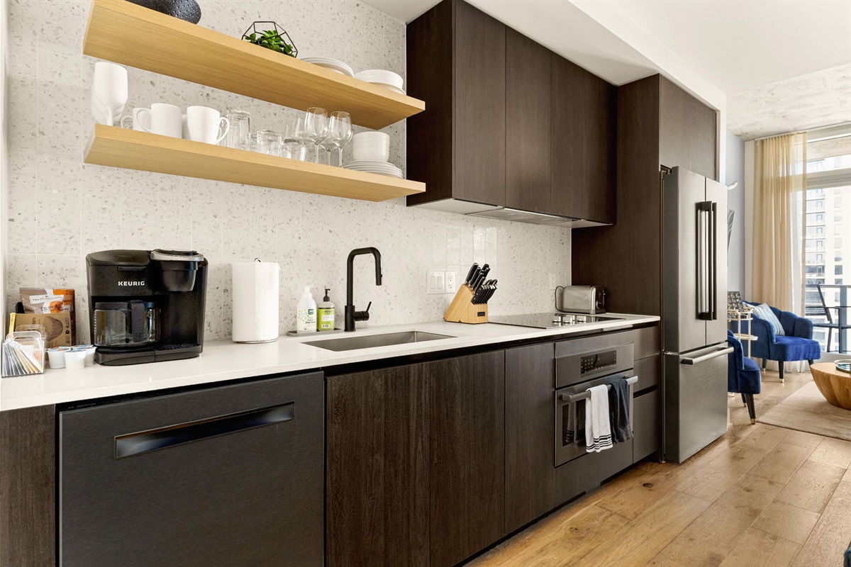 A stylish and functional kitchen featuring dark wood cabinetry, light-colored countertops, and a speckled white backsplash. The space is well-equipped with a full-sized stainless steel refrigerator, built-in oven, dishwasher, and modern appliances like a K