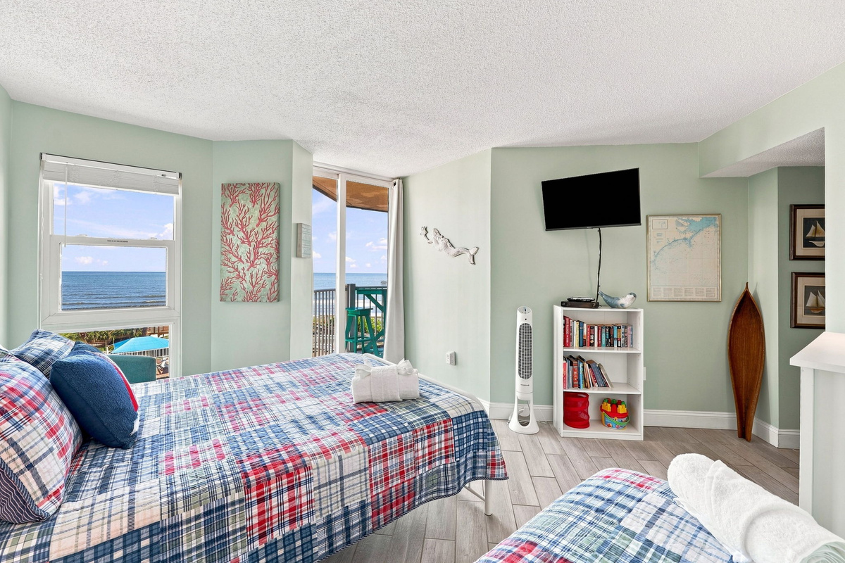 Guest bedroom featuring two cozy beds, vibrant coastal decor, and a sliding glass door opening directly to the oceanfront balcony.
