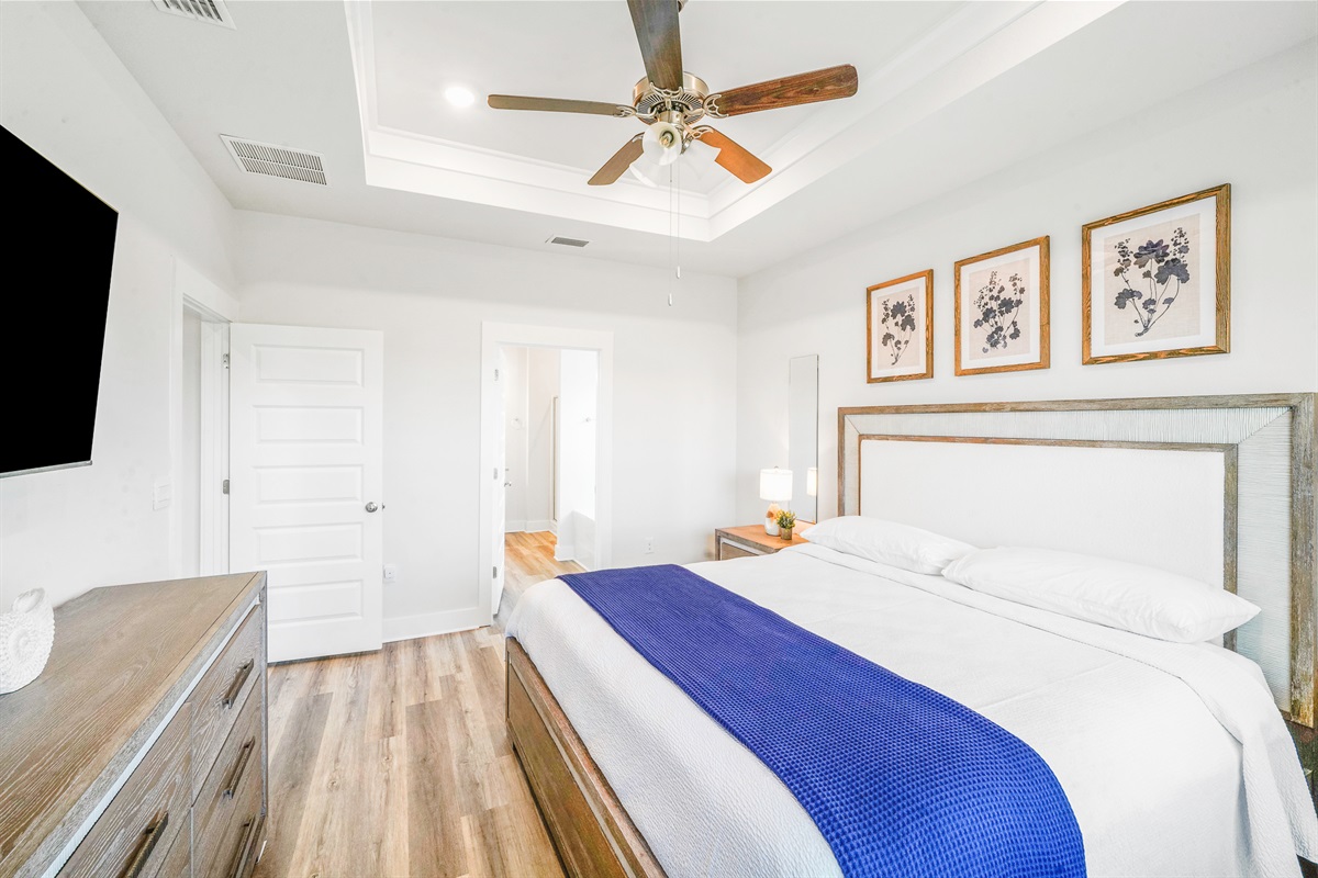 Primary Suite: Modern king bedroom featuring an en suite bathroom, plush linens, ceiling fan, elegant bedside lamps, dresser, and Smart TV.