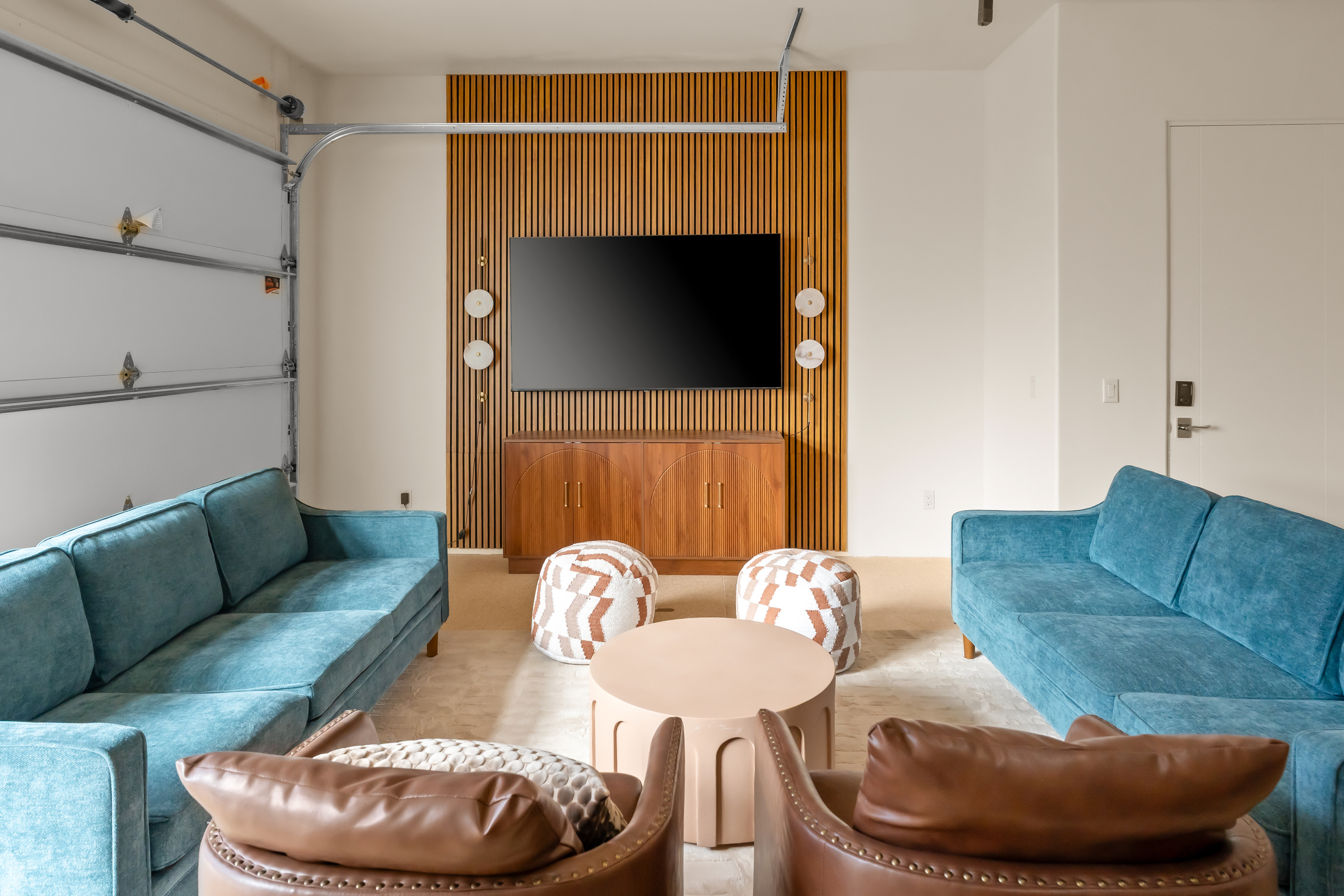 Movie night ready: deep teal sofas, leather armchairs, cozy poufs, and a large flat-screen 75" TV framed by modern timber paneling.