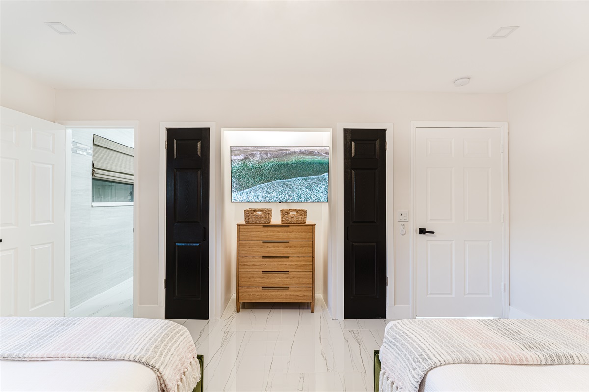 Bright coastal suite with two queen beds, private ensuite bath, smart TV, blackout bamboo shades, and a rattan ceiling fan—ideal for families or shared stays.
