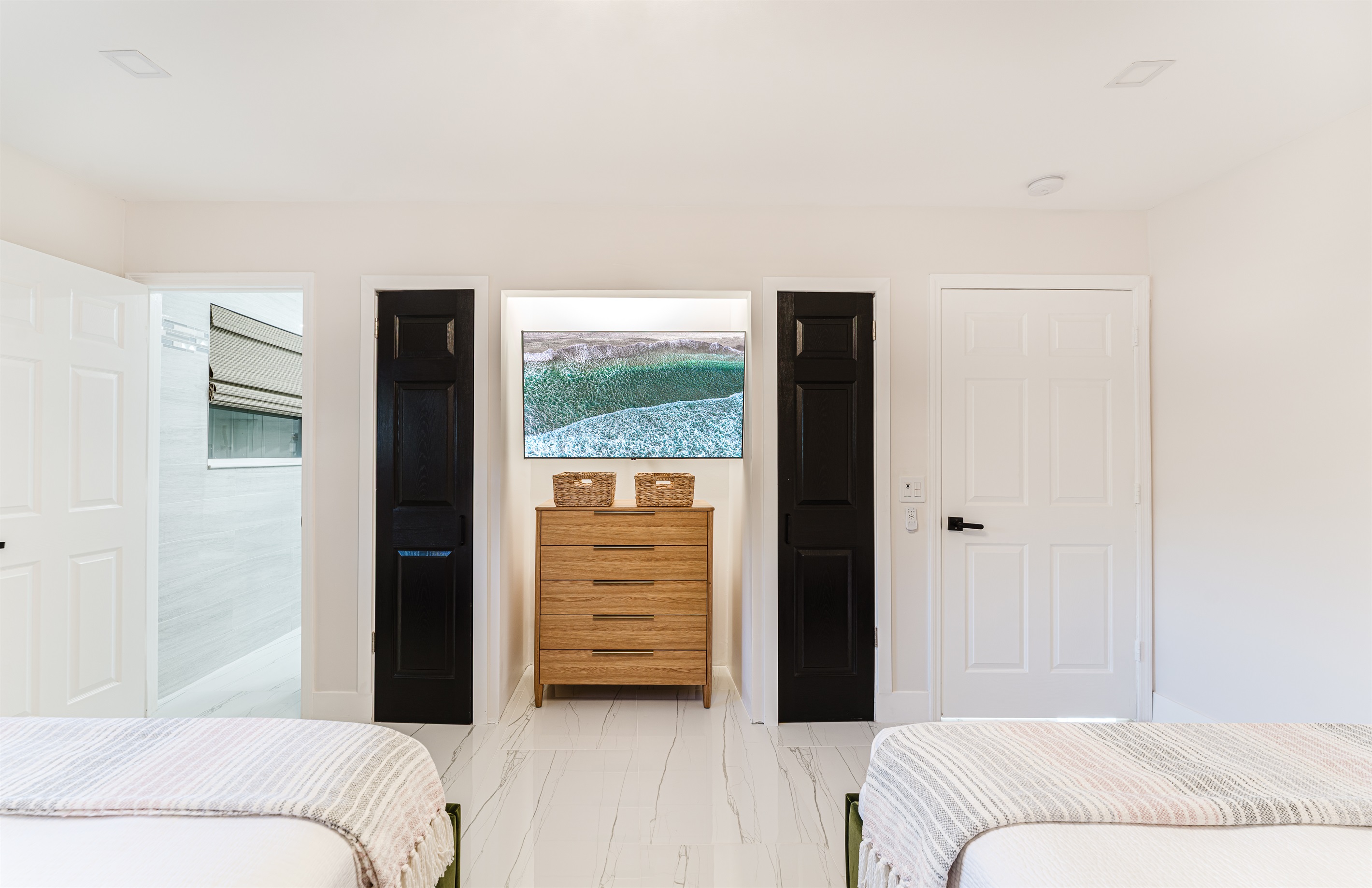Bright coastal suite with two queen beds, private ensuite bath, smart TV, blackout bamboo shades, and a rattan ceiling fan—ideal for families or shared stays.