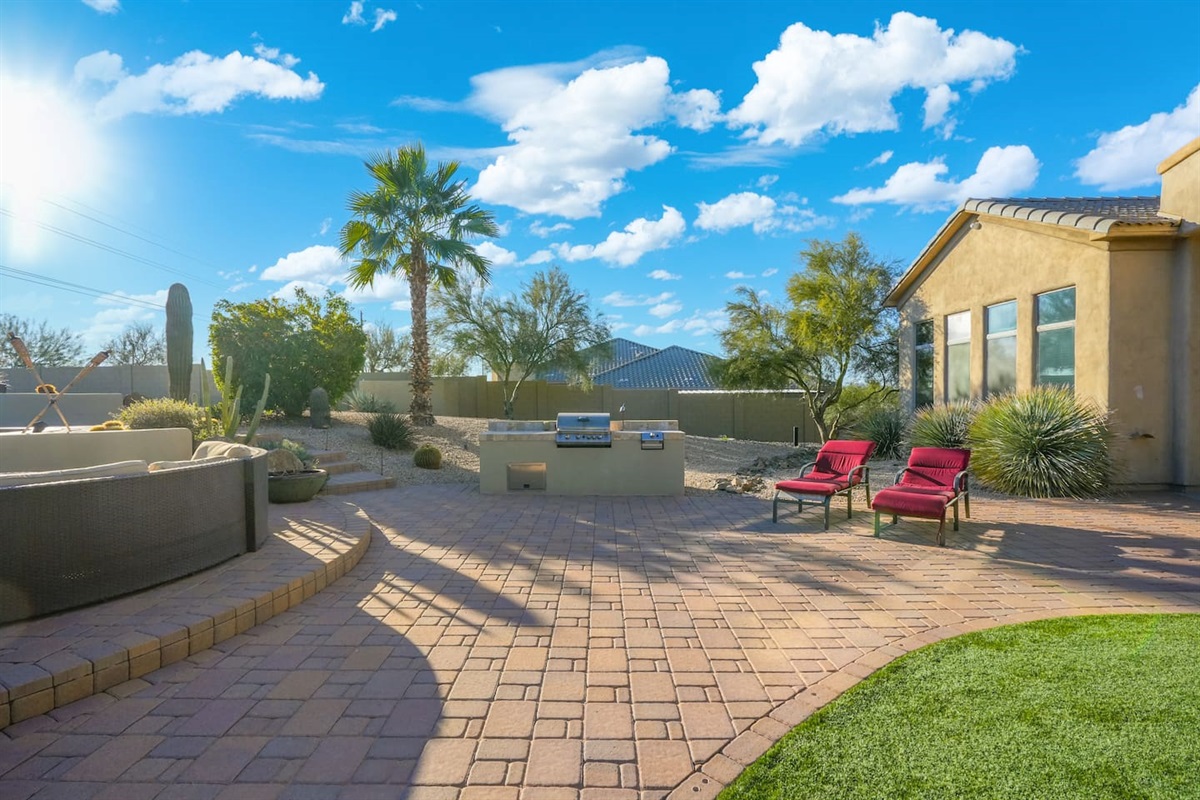 Wide patio view featuring the built-in BBQ, curved fire pit seating, and red loungers—perfect for entertaining and enjoying sunny desert days.
