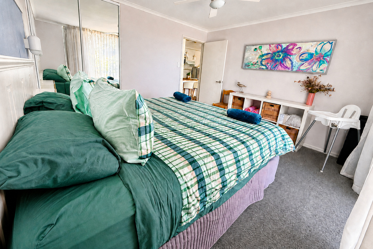 This light-filled bedroom at Josh’s Sea Breeze includes mirrored robes, a ceiling fan, bedside lighting and soft green bedding for an easy coastal stay.
