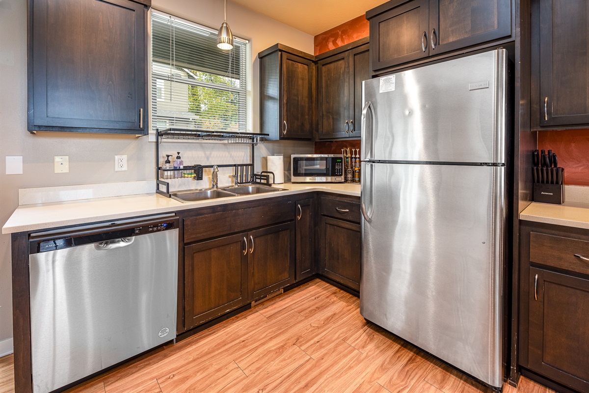 A modern kitchen featuring dark wood cabinets, a stainless steel refrigerator, a silver dishwasher, a double sink with a sleek faucet, a microwave, and a small shelf with cleaning supplies, all set against a warm-toned wall.