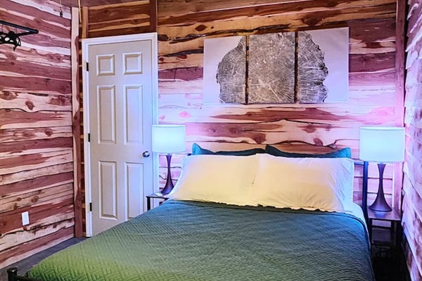 This cozy bedroom features warm cedar walls, a braided rug, and a plush queen bed with a deep green quilt. Soft lighting and a striking tree-ring wall art create a calming, nature-inspired space perfect for unwinding after a day outdoors.