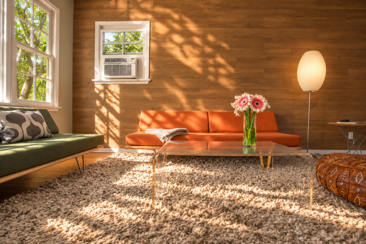 The sunlit living room combines an orange sofa and green seating with a clear acrylic coffee table, textured rug and leather pouf. Leaf-shaped shadows play across the wood feature wall, and a glowing floor lamp adds warmth beside the sofa.