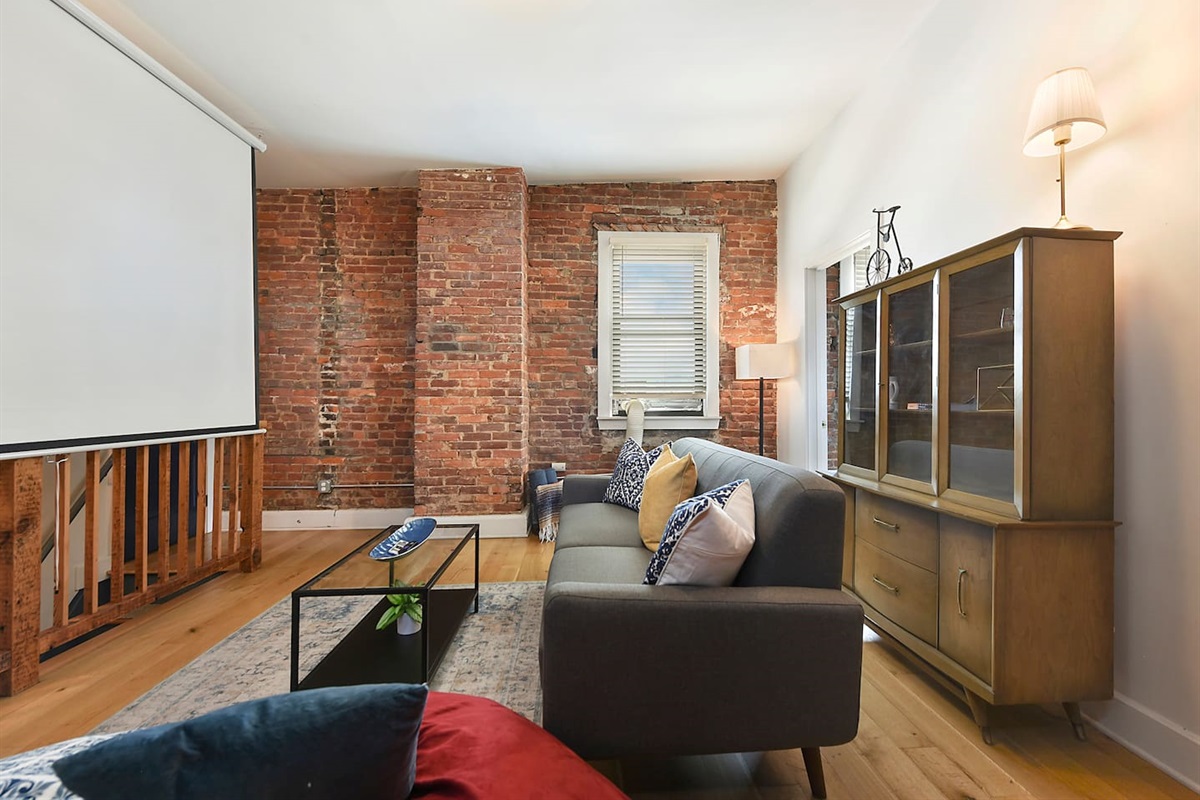 Kick back in this inviting living room with a cushioned couch, bean bag, and a center coffee table, all set against a warm brick wall and a plush rug—plus a projector screen for the best entertainment.