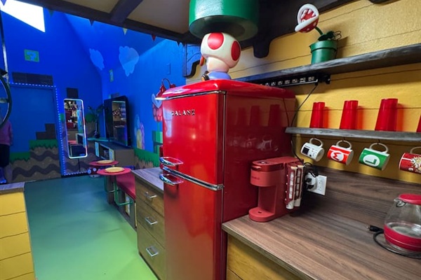 Whimsical full-yet-compact kitchen at Mario's Mini Mansion, Clermont, FL! Bright red retro-style fridge, green flooring, yellow walls with Mario shelves, and Toad-inspired decor make post-theme park meal prep an adventure for kids and adults alike.