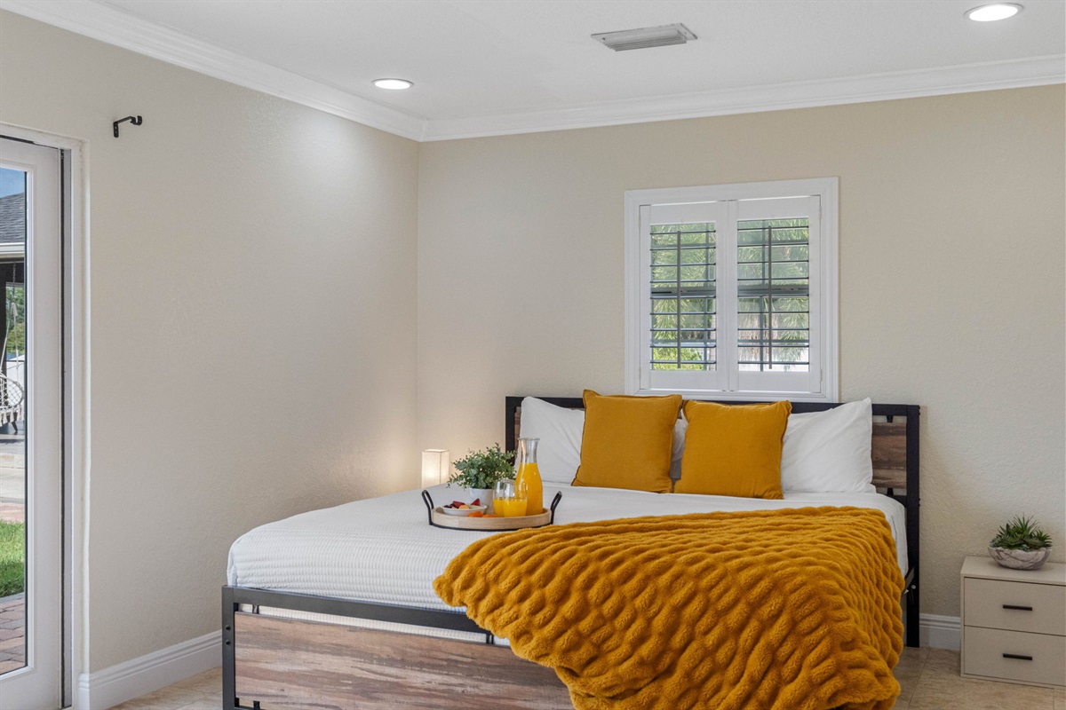 A modern bed dressed in crisp white and rich golden hues sits beside windows framing tranquil palm and water views.