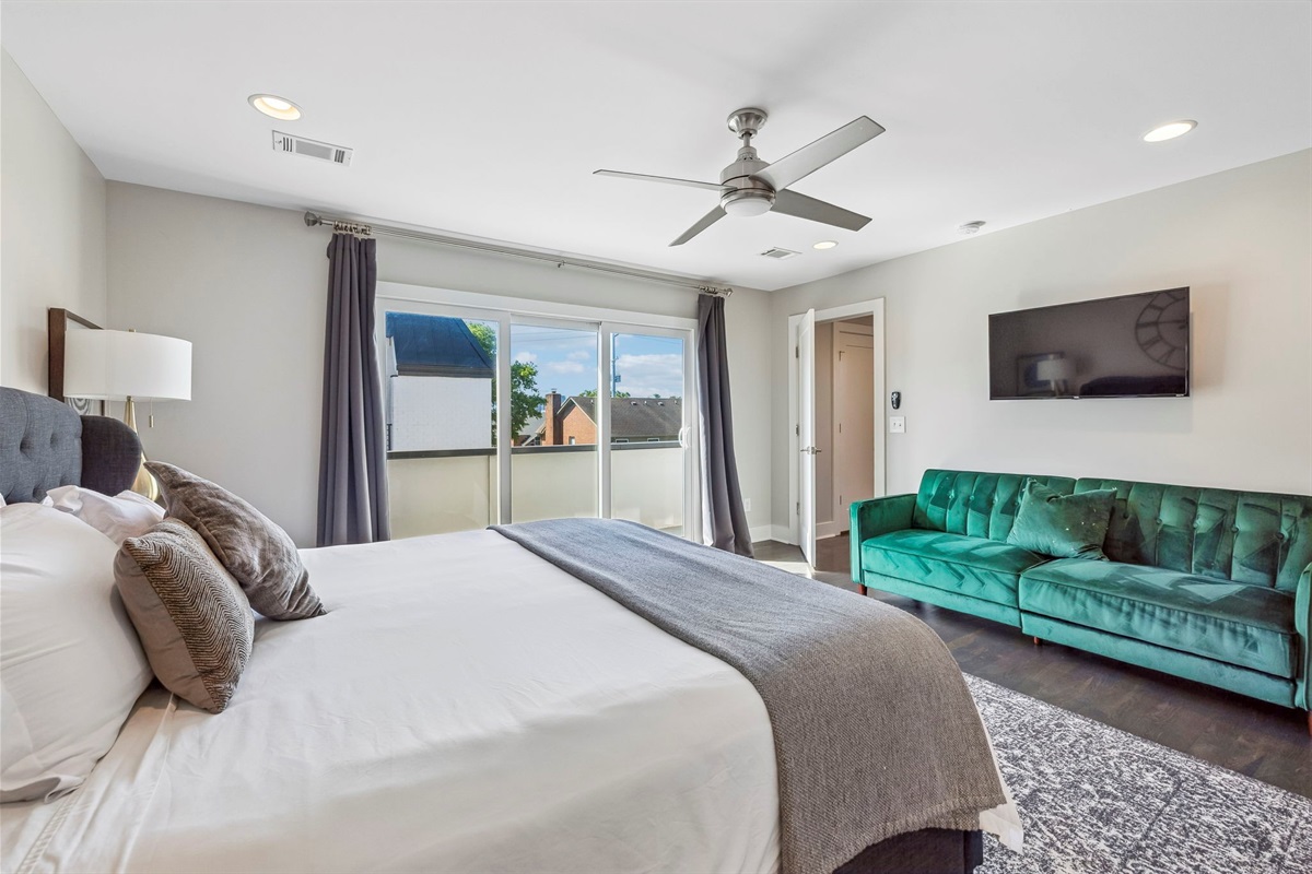 Emerald green tufted sofa adds a vibrant pop of color to this spacious bedroom lounge - Wall mounted TV and large windows make this a great spot for relaxing or streaming - Sophisticated black out drapes and detailed rug complete the room&rsquo;s upscale feel