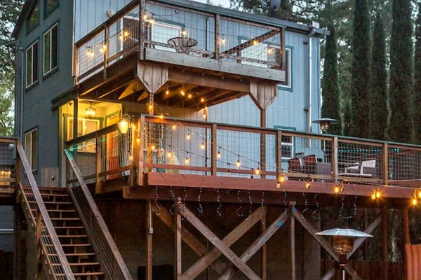 - Elevated two story home with wraparound deck and string lights for charm  
- Outdoor seating with fire pit creates a cozy gathering space at night  
- Surrounded by tall pines for a peaceful and private retreat experience  
