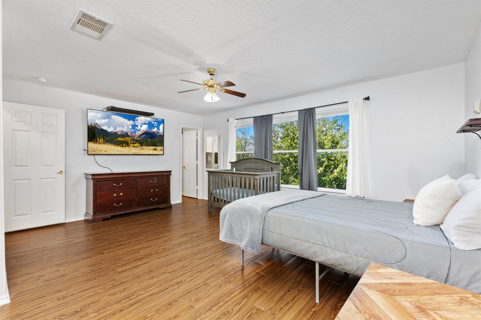 King bedroom with wall-mounted Smart TV, ceiling fan, natural light, gray bedding, and room-darkening shades. Clothing storage and extra pillows included.