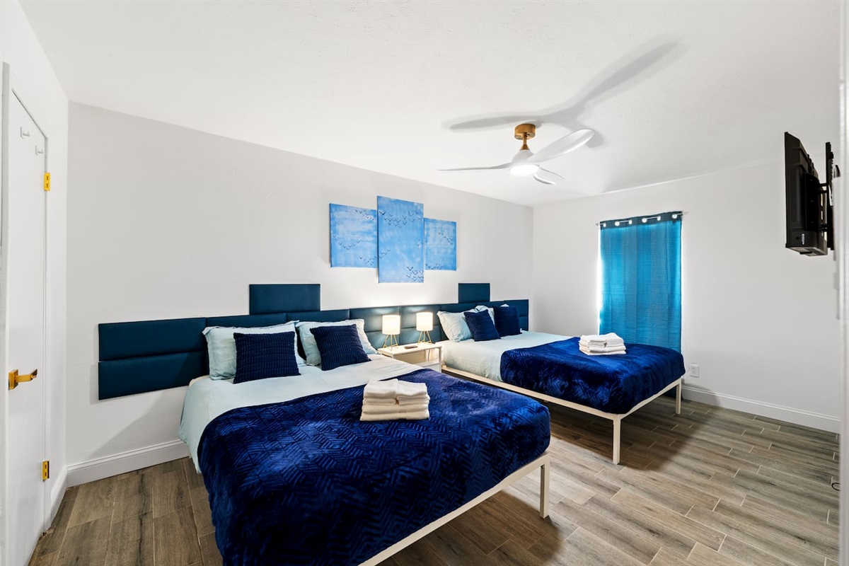 Spacious bedroom with two plush queen beds, blackout curtains, and a smart TV—ideal for families or groups to unwind in comfort.