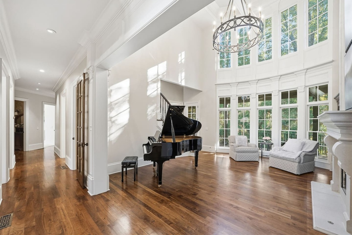 Beautiful entry sitting room with a grand piano, warm fireplace, tall windows, and two comfortable chairs. A bright, serene spot to unwind or enjoy quiet moments.