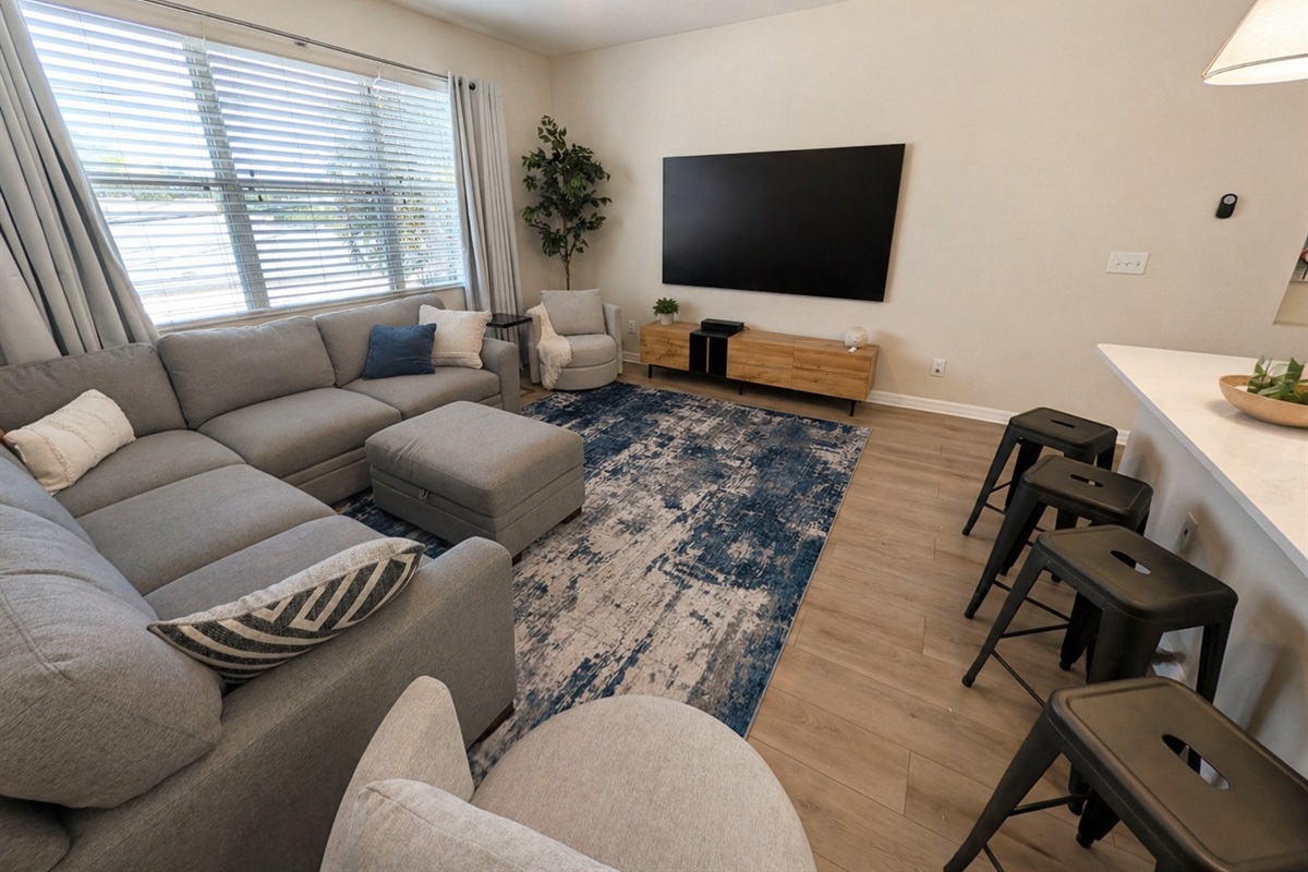 Bright, open living room with a large sectional sofa and ottoman—perfect for relaxing or gathering. Enjoy natural light from oversized windows, a big-screen TV, and easy access to the kitchen with bar seating for casual dining.