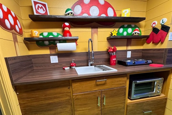Fully equipped Nintendo-inspired kitchen! Warm wood cabinets, dark countertops, built-in microwave, sink, and vibrant Super Mario mushroom shelves stocked with fun details—perfect for family meals at Mario's Mini Mansion.