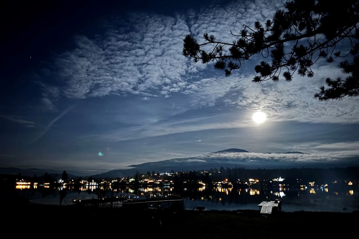 A glowing moon rises above a clouded sky, casting light over the lake as shoreline lights shimmer on gentle ripples. The horizon softens into a pale blue, creating a serene, moody evening where water, clouds, and moonlight meet.