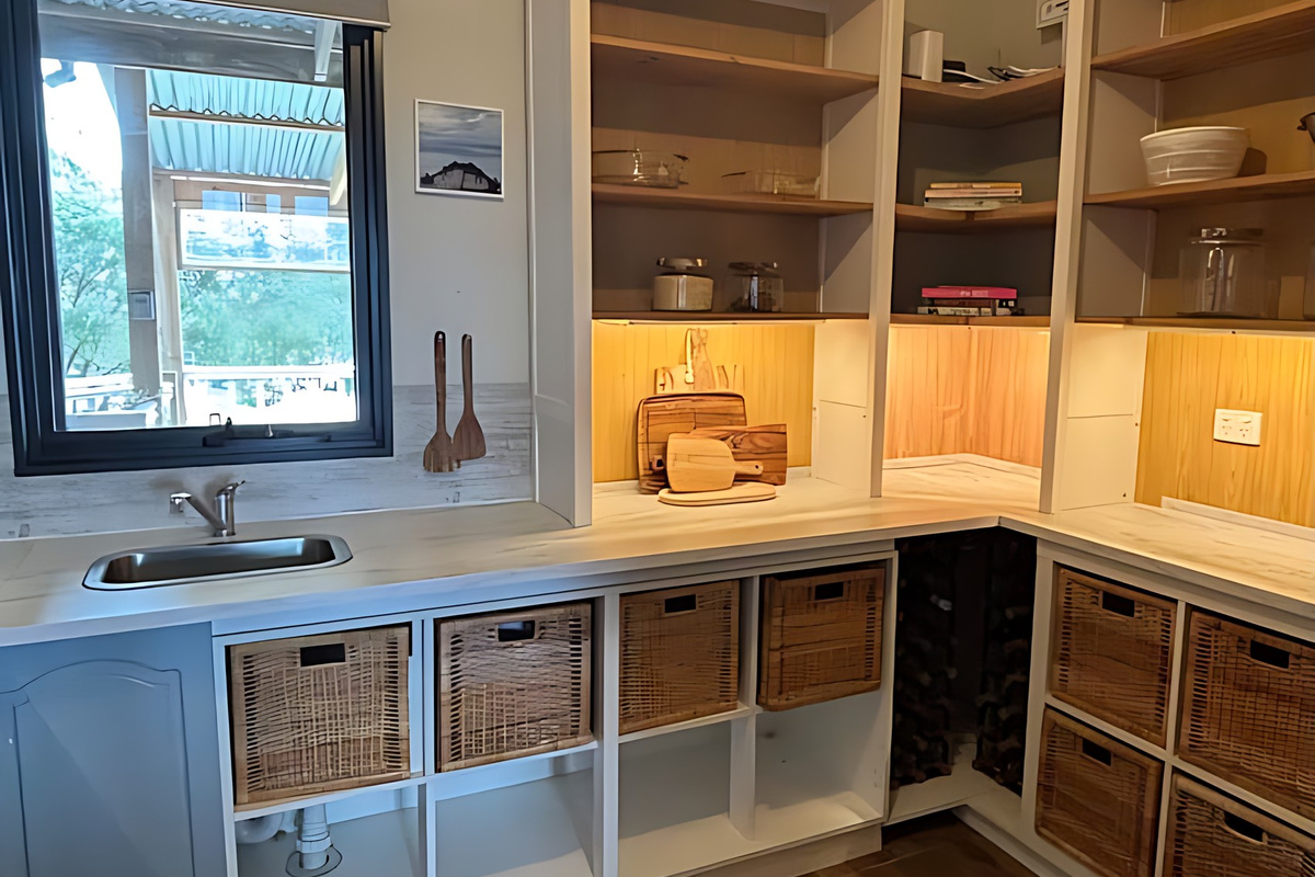 The scullery at Lake Views & Coastal Escape adds a second sink under the window, open timber shelving, wicker storage baskets and a built-in wine rack, all a few steps from the main kitchen.