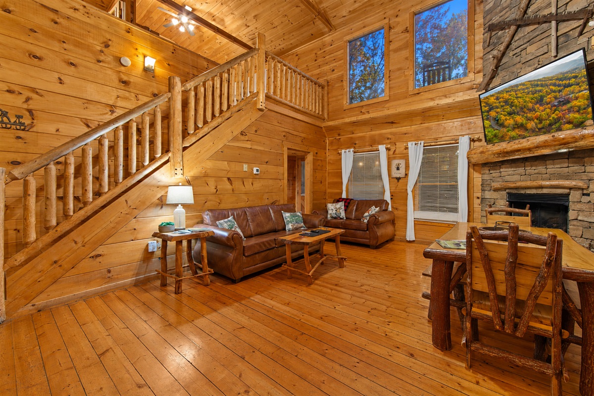 This open-concept cabin living area boasts cozy leather couches, a rustic wooden staircase, and a fully equipped kitchen with black appliances, all set within a warm, wood-paneled interior.