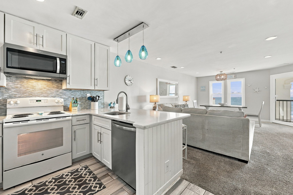 Open-plan kitchen featuring white shaker cabinets, stainless steel appliances, mosaic tile backsplash, blue pendant light fixtures, and a beadboard island base opening to the living and dining areas.   