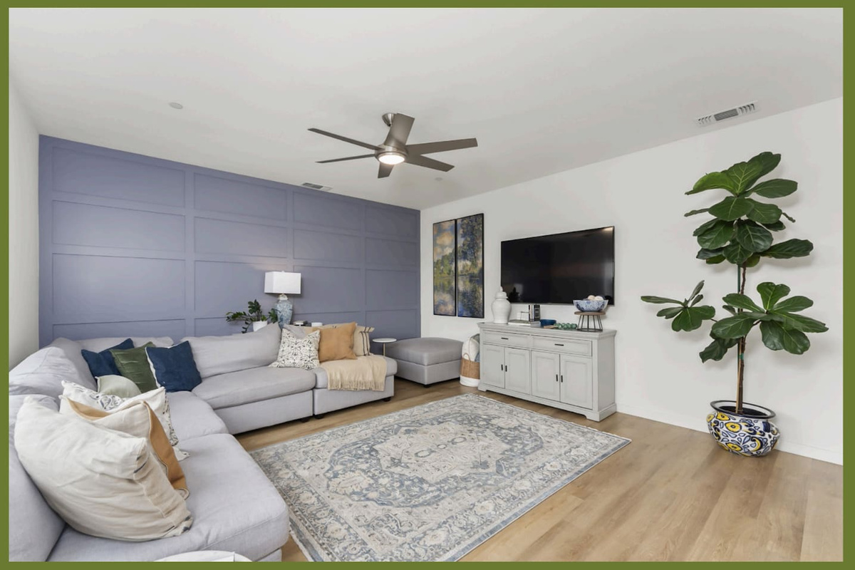 Bright living room with sectional seating, wall-mounted TV, ceiling fan, and a large window overlooking the backyard.